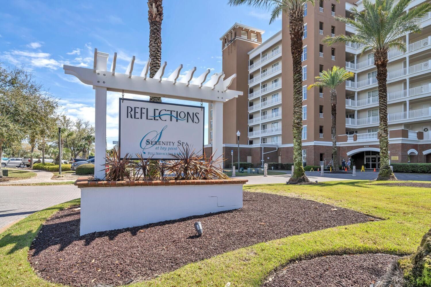 A hotel entrance with a sign for 'Reflections Serenity Spa' at bay point, featuring a modern building with balconies and palm trees.