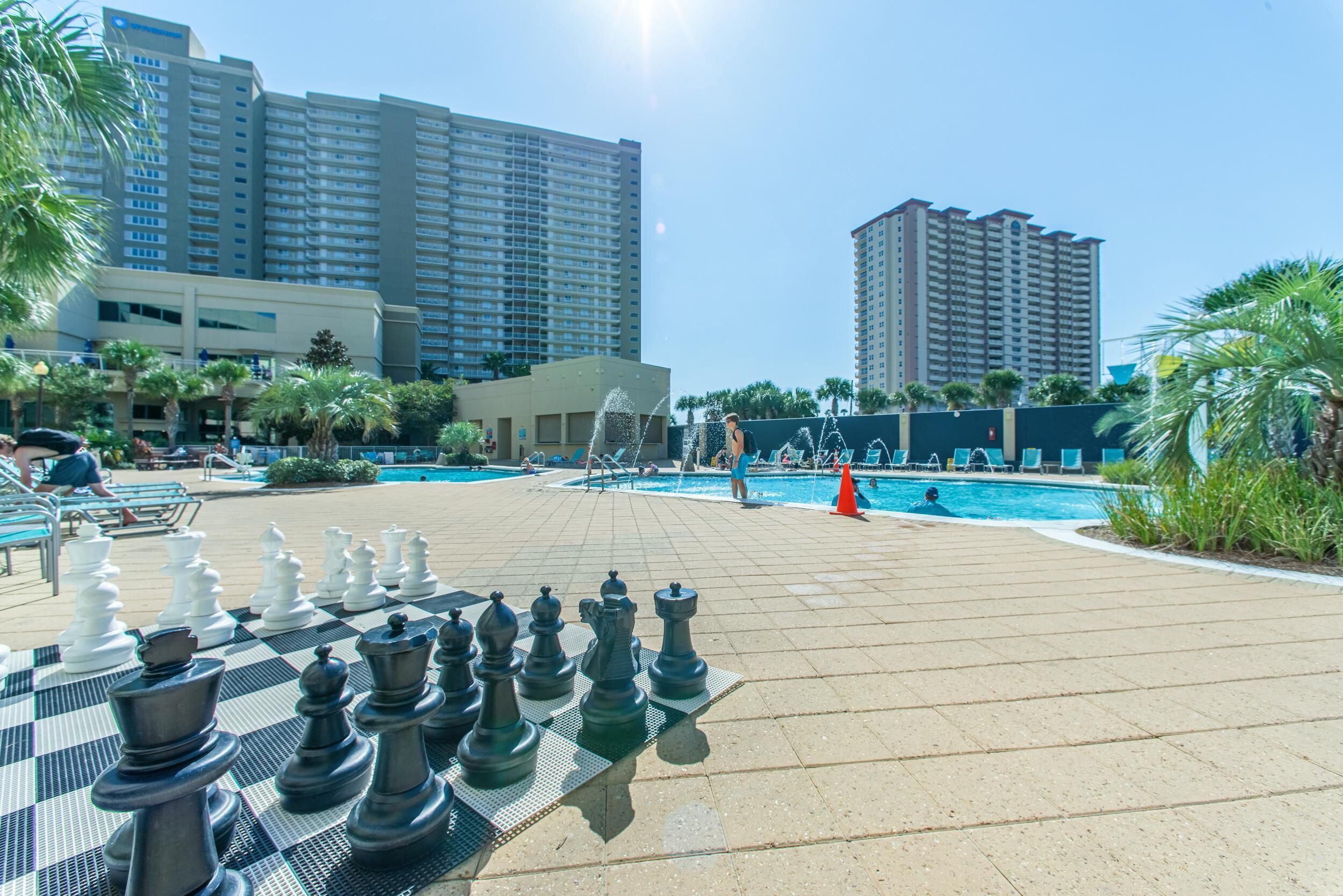 A pool area with chess pieces set up on a table, surrounded by palm trees and high-rise buildings in the background.