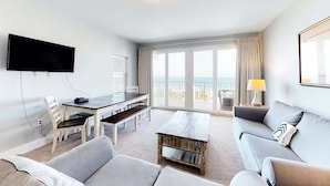 Una habitación de hotel moderna con una televisión de pantalla plana, una mesa de comedor con sillas, un sofá, una mesa baja de madera y un amplio ventanal con vista al océano.