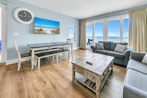 A spacious living room with a wooden floor, a dining table with chairs, a gray sofa, and a large window offering a view of the ocean. - Laketown Wharf 801 (Panama City Beach)