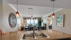A kitchen with granite countertops, a double sink, and a dishwasher. There is a dining area with a table and chairs, a ceiling fan, and pendant lights. A decorative metal wall piece and a framed picture are visible. - Marisol 204 (Panama City Beach)