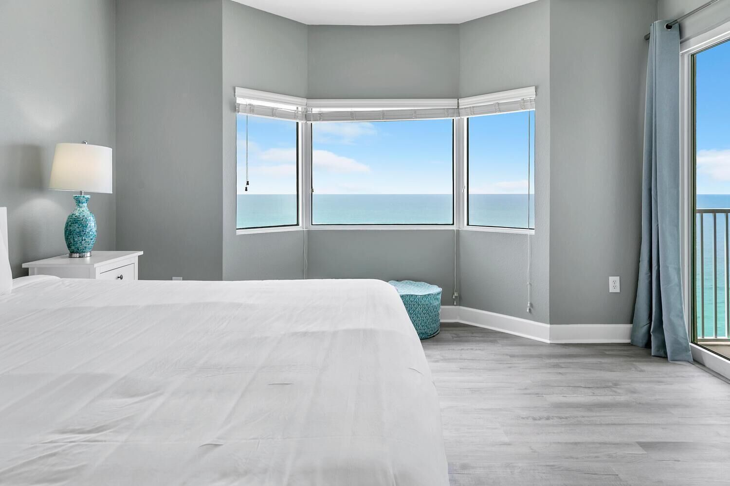 A bedroom with a large bed, a nightstand, a lamp, a blue vase, a window with a view of the ocean, and a corner window with a view of the sky.