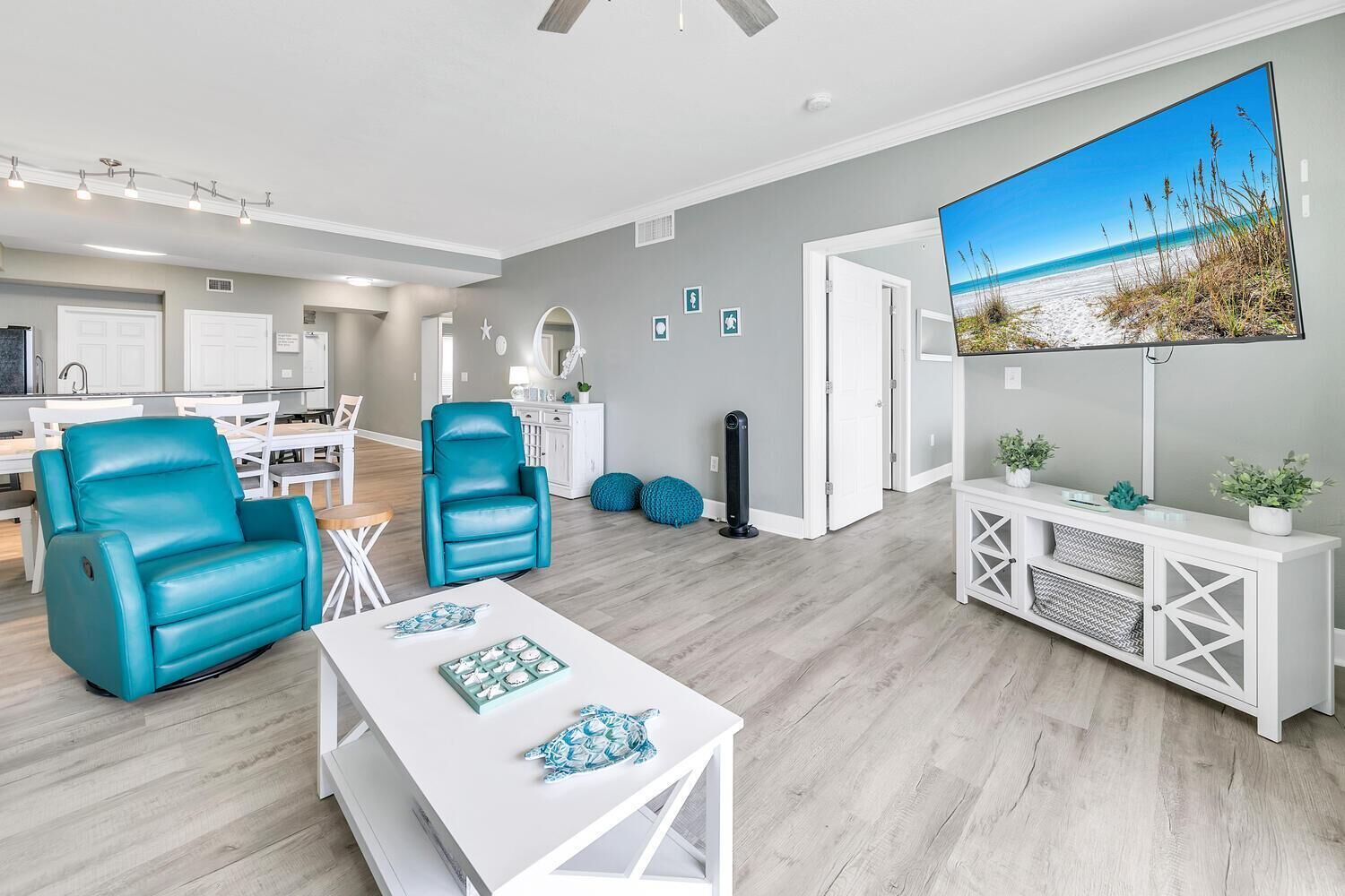 A modern living room with teal leather chairs, a white coffee table with decorative items, a large flat-screen TV on a white console, and a view of the beach through a large window.