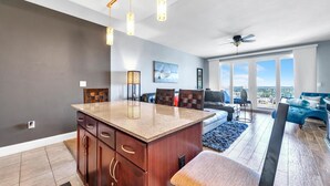 A modern kitchen with a central island, a dining area with a blue sofa, and a view of the city through large windows. - Laketown Wharf 1829 (Panama City Beach)