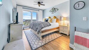 A bedroom with a bed, a desk, a chair, a ceiling fan, a window, and a wall clock. - Laketown Wharf 1829 (Panama City Beach)