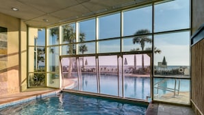 Una piscina interna con pareti di vetro trasparenti, pavimento piastrellato e vista su palme e un edificio in lontananza.