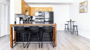 Eine moderne Küche mit Holzausstattung, einem Edelstahlkühlschrank, einer Mikrowelle und einem Gasherd. Drei schwarze Barhocker und ein kleiner runder Tisch mit zwei Hockern ergänzen das Bild.