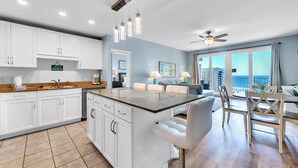 A modern kitchen with white cabinets, a granite countertop, and a bar stool. There is a dining area with a table and chairs, and a view of the ocean through large windows. - Laketown Wharf 827 - Life's A Beach (Panama City Beach)