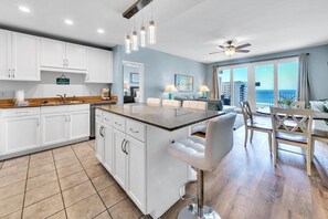 A modern kitchen with white cabinets, a granite countertop, and a bar stool. There is a dining area with a table and chairs, and a view of the ocean through large windows. - Laketown Wharf 827 - Life's A Beach (Panama City Beach)