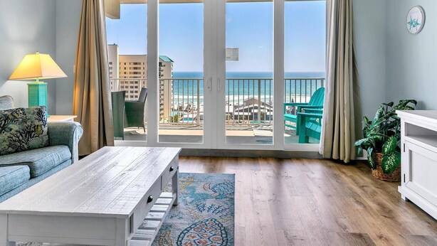A room with a large sliding glass door leading to a balcony with a view of the beach and ocean. There is a sofa, a coffee table, and a potted plant. - Laketown Wharf 827 - Life's A Beach (Panama City Beach)
