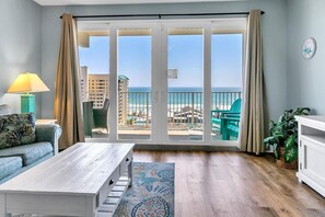 Habitación con un amplio ventanal corredero que da a un balcón con vistas a la playa y al océano. Se observa un sofá, una mesita de centro y una planta en maceta.