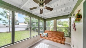 Ein Wintergarten mit großen Fenstern, einem Deckenventilator, einer hölzernen Bank und einem Hängekorbsessel.