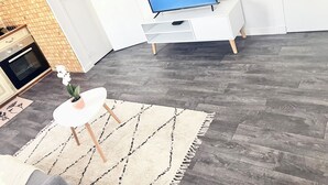 Moderni olohuone, jossa on litteänäyttöinen televisio, valkoinen sohva, pieni valkoinen pöytä, jossa on kasvi, ja kuviollinen matto.