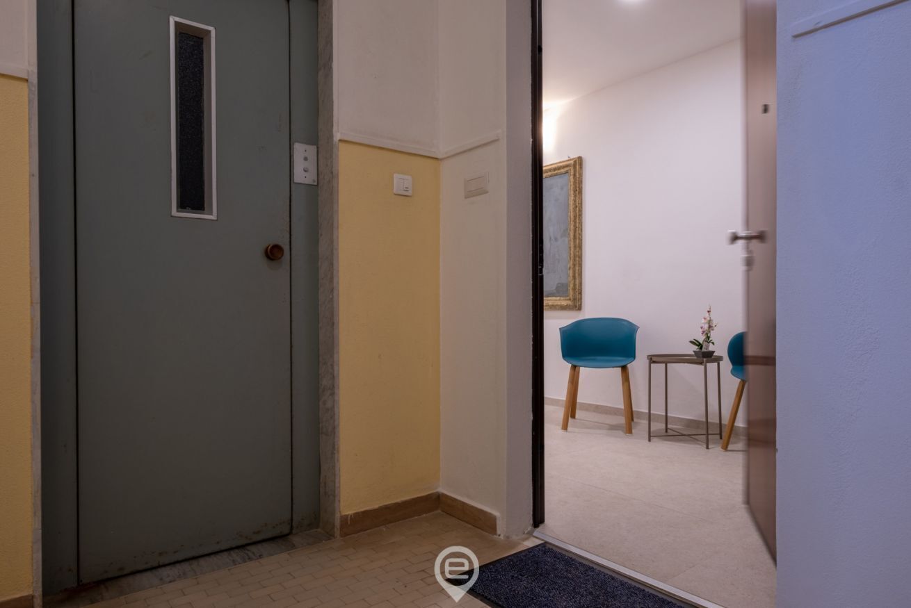 Un pasillo con una puerta gris, un ventanal pequeño y una silla azul cerca de una mesa con un florero.