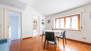 A spacious room with a wooden floor, a round dining table with chairs, a small plant on the table, and a window with handwritten notes.