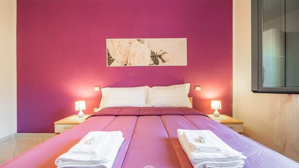 A hotel room with a large bed, two bedside tables with lamps, a painting on the wall, and towels on the bed. - Dahlia Apartment (Cabras)