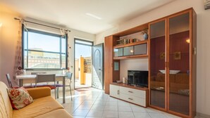 A living room with a sofa, TV stand, and a sliding glass door leading to a balcony. - Dahlia Apartment (Cabras)