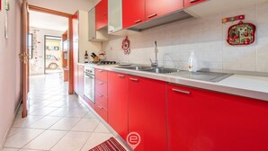 A kitchen with red cabinets, a stainless steel sink, and a tiled backsplash. - Dahlia Apartment (Cabras)