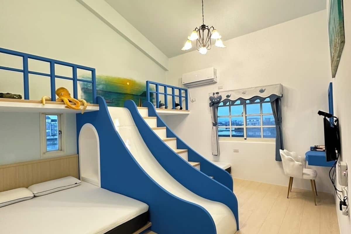 A room with a bunk bed, a blue and white slide, a desk with a chair, and a window with a view.