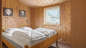Ein Zimmer mit Holzwänden, einem Bett, einem Fenster mit Jalousien und zwei gerahmten Bildern an der Wand.