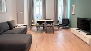 Ett modernt vardagsrum med en grå soffa, ett matbord med fyra stolar, en tv på ett tv-bord och stora fönster med gardiner.