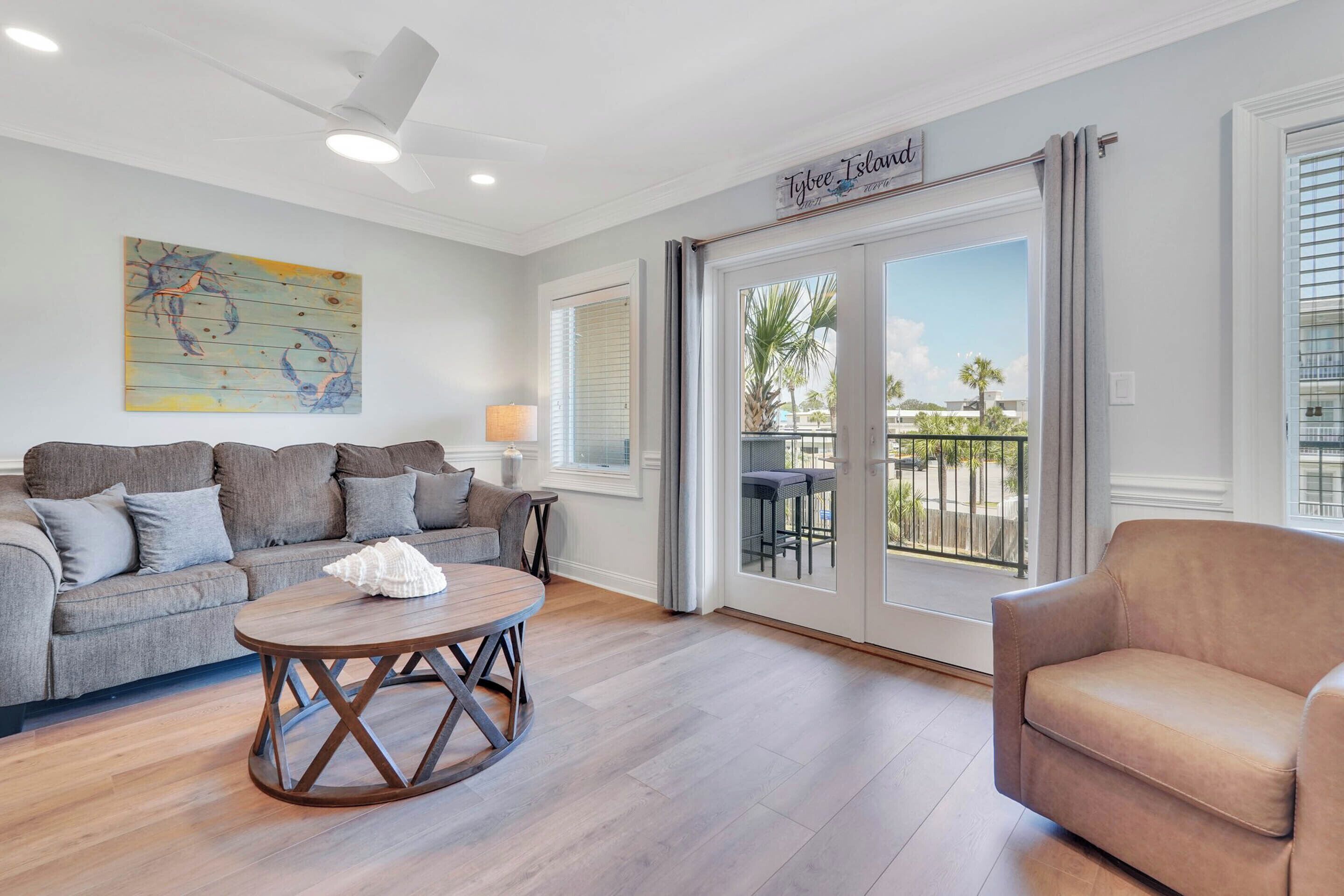 Ein Wohnzimmer mit grauem Sofa, einem runden Holztisch und einem beigen Sessel. Ein großes Fenster bietet Blick auf Palmen und einen Balkon.