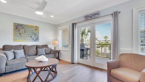 A living room with a grey sofa, a round wooden coffee table, and a beige armchair. There is a large window with a view of palm trees and a balcony.