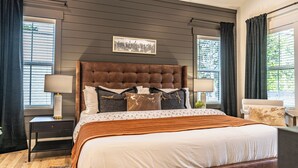 A bedroom with a large bed, dark wood paneling on the walls, two bedside tables with lamps, and a window with curtains. - The Cowboy (Southern Pines)