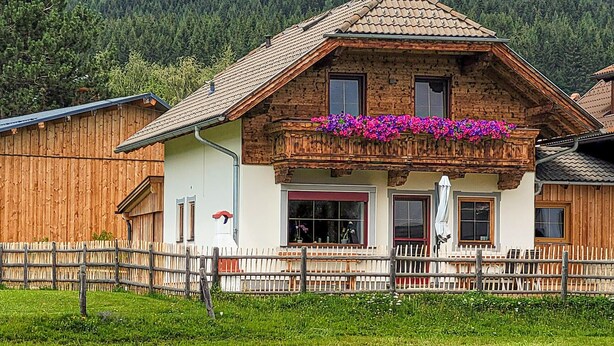 A traditional chalet with a wooden facade, a balcony with flowers, and a view of a forested mountain in the background. - Beautiful home in Mariapfarr with WiFi (Mariapfarr)