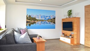A modern living room with a large flat-screen TV mounted on a wooden wall unit, a grey sofa with cushions, a small wooden coffee table, and a large framed landscape painting of a mountain lake. - Beautiful home in Mariapfarr with WiFi (Mariapfarr)
