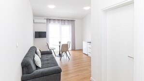 A modern living room with a grey sofa, a small round table, and two chairs. There is a TV mounted on the wall, a window with curtains, and a white door. - 1 bedroom nice apartment in Kastel Kambelovac (Kastel Kambelovac)