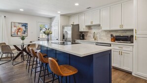 A modern kitchen with white cabinets, a large island with bar stools, a refrigerator, and a microwave. - Feel at Home! New Renovated House By *Grandscapes* (The Colony)