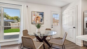 A dining area with a glass table and wicker chairs, a painting on the wall, and a view of a backyard through a sliding door. - Feel at Home! New Renovated House By *Grandscapes* (The Colony)