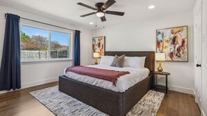 A bedroom with a large bed, a ceiling fan, two bedside lamps, and artwork on the walls. - Feel at Home! New Renovated House By *Grandscapes* (The Colony)