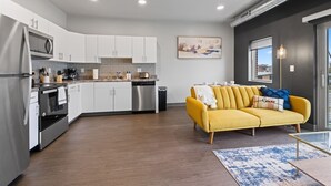 Une cuisine moderne avec des meubles blancs, des appareils en acier inoxydable et un canapé jaune dans un salon agrémenté d’un tapis à motifs bleus.