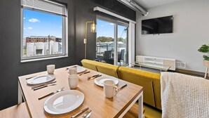 Un salon moderne comprenant un coin repas, un canapé, un téléviseur fixé au mur et une fenêtre avec des stores.
