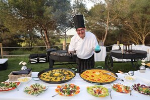 A chef at an outdoor event with large pans of food and various dishes on a table.