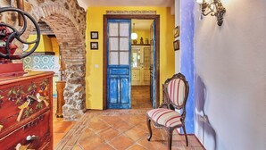 Habitació amb paret de pedra, una porta blava, un aparador de fusta amb motius de fruita i una cadira rayada.