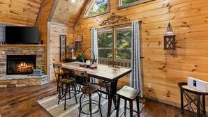 A cozy wooden cabin interior with a stone fireplace, a dining table with chairs, and a TV mounted above the fireplace.