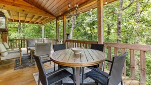 A wooden deck with a round table and chairs, surrounded by a wooden railing, overlooking a forested area.