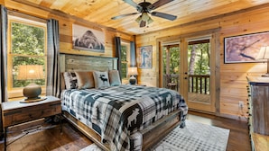 A wooden cabin bedroom with a bed, a nightstand, a ceiling fan, and large windows.
