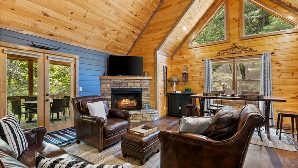 A cozy log cabin interior with a stone fireplace, leather furniture, and large windows offering views of the outdoors.