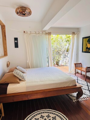 A bedroom with a wooden bed, white curtains, a view of greenery outside, and a wooden bench by the window. - Villa Samaé, Tropical charm in the south-west of Reunion Island (Les Avirons)