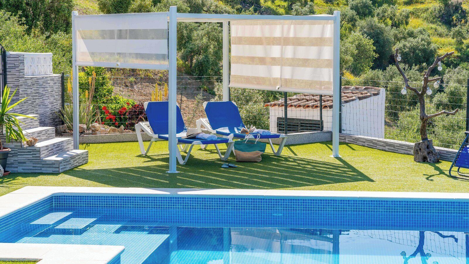 A swimming pool with blue tiles, two blue lounge chairs, a white pergola, and a garden with plants and a tree.