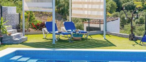 A swimming pool with blue tiles, two blue lounge chairs, a white pergola, and a garden with plants and a tree.