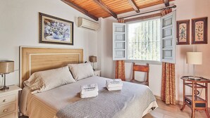 A bedroom with a wooden headboard, a bed with a patterned bedspread, a nightstand with a lamp, a chair, and a window with curtains.