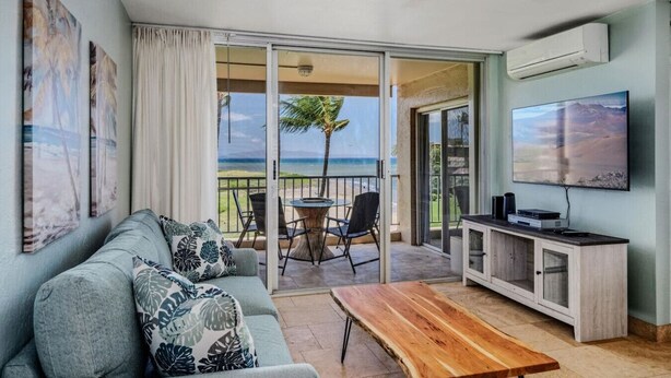 A living room with a sofa, coffee table, TV, and a balcony with a view of the beach. - Amazing Oceanfront Views! MS420 (Kihei)
