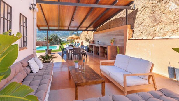 An outdoor patio with a wooden table, a sofa, and a dining area with chairs and a table. There is a pool in the background. - Villa Los Ruedos Riogordo by Ruralidays (Málaga)