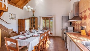 Un coin repas avec une longue table dressée pour le repas, des chaises en bois, un buffet avec des portes en verre et un espace cuisine équipé d’une cuisinière et d’un four à micro-ondes.