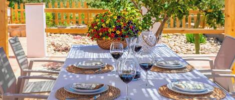 Un comedor al aire libre con una mesa dispuesta para cuatro personas, rodeada de sillas, con una valla de madera y follaje al fondo.